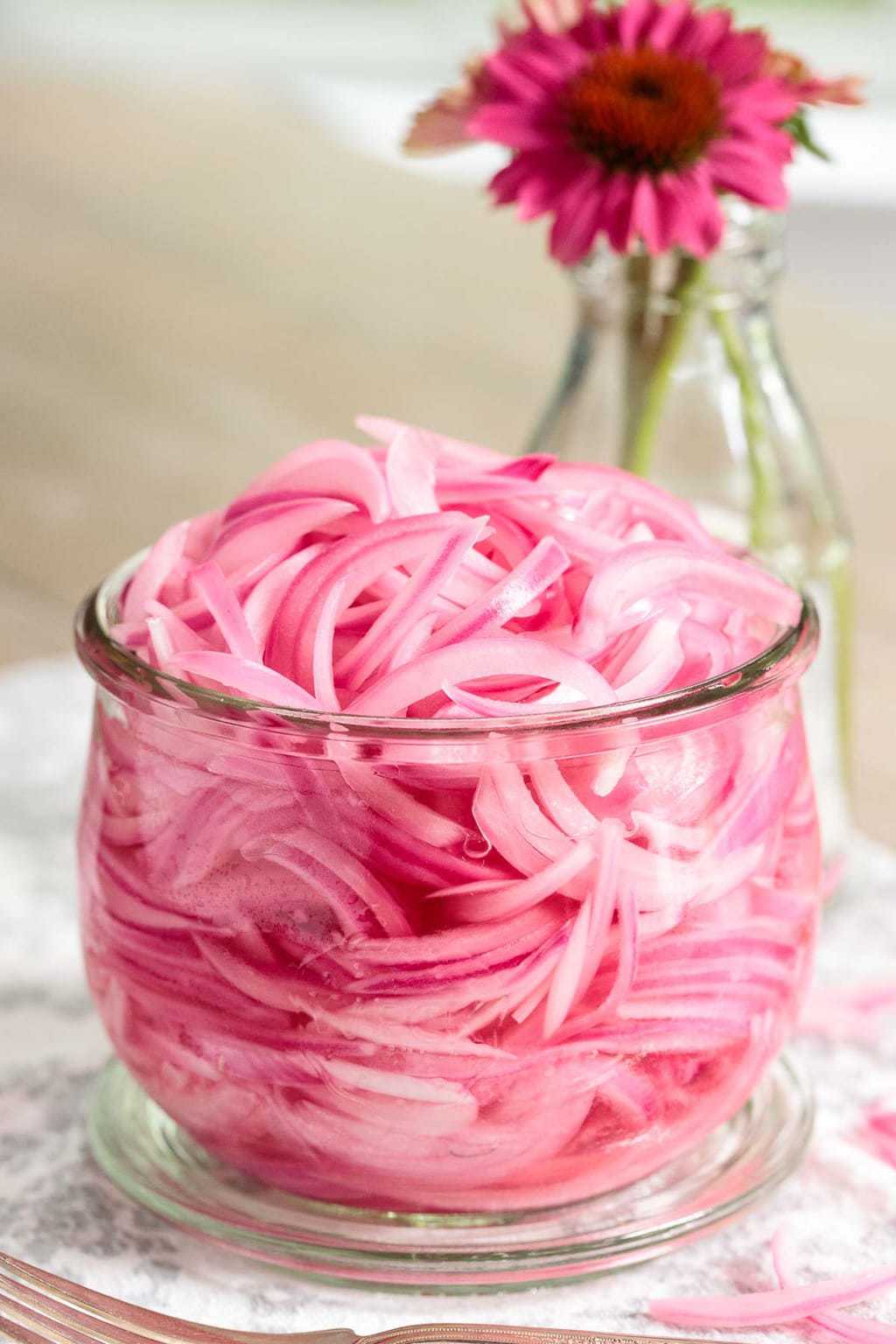 Photo of a glass jar of Pickled Red Onions on a table with a coneflower in a vase in the background.