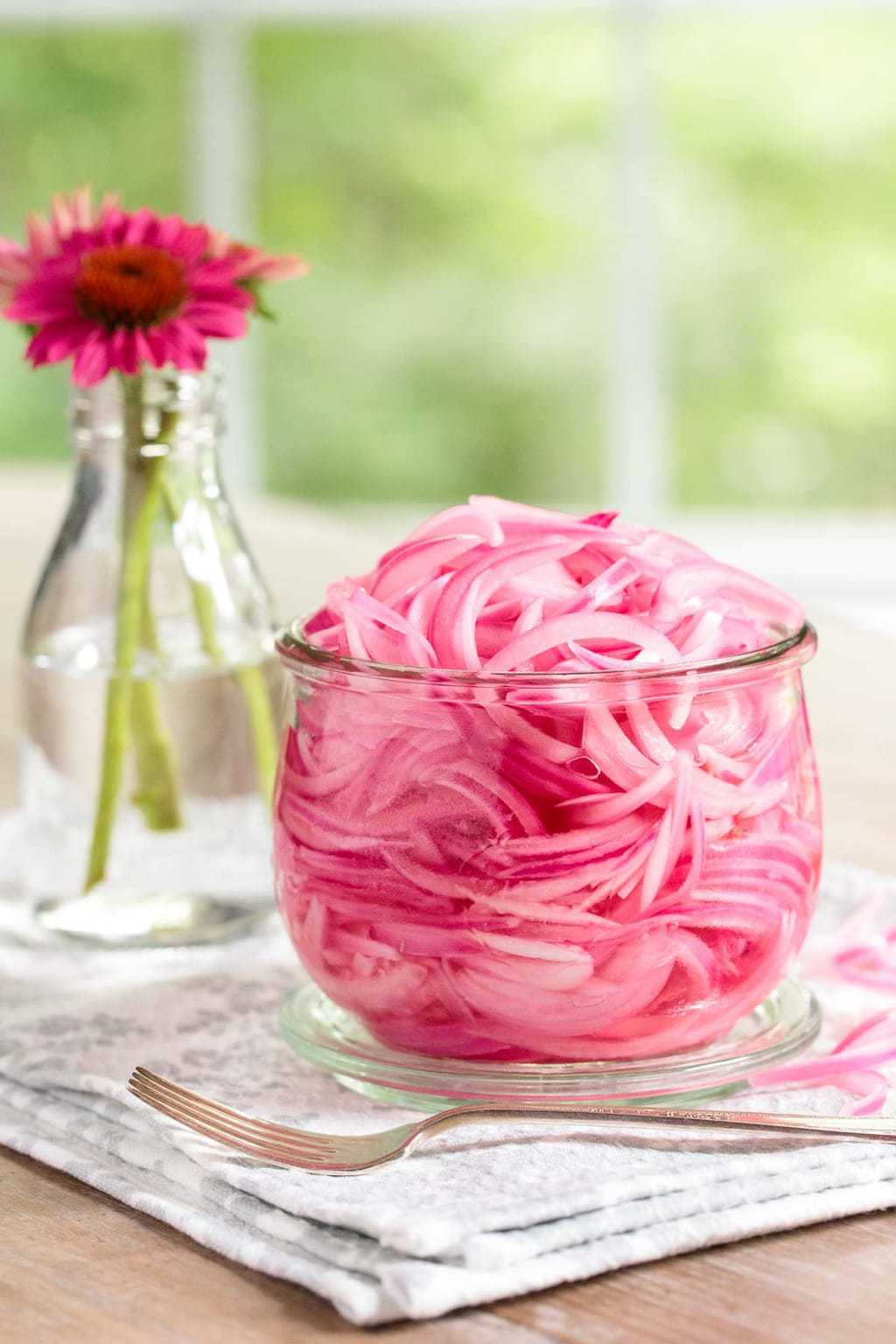 Photo of a batch of Easy Pickled Red Onions in a glass jar on a dining room table with a vase of cone flowers in the background.