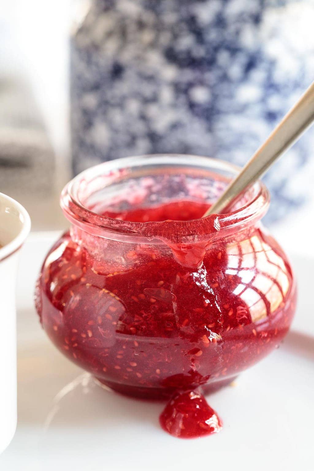 Photo of a jar of Easy Raspberry Freezer Jam with jam drizzling down the front and a blue and white porcelain jar in the background.