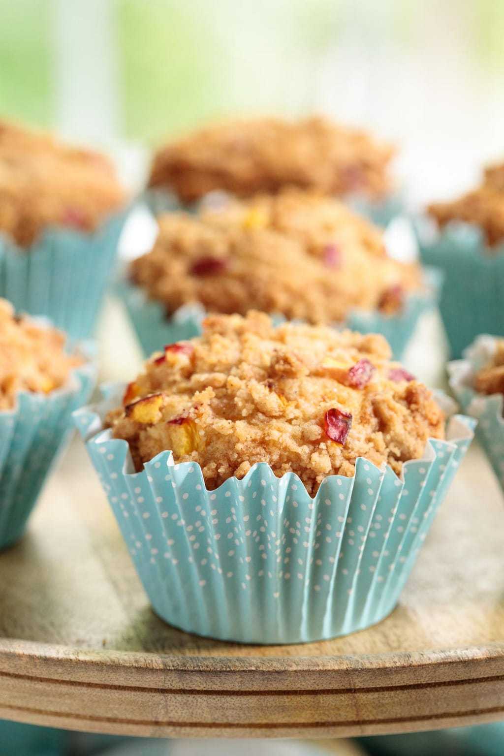 Vertical closeup photo of a batch of Peach Crumble Muffins in turquoise muffin liners on a wood serving plate.