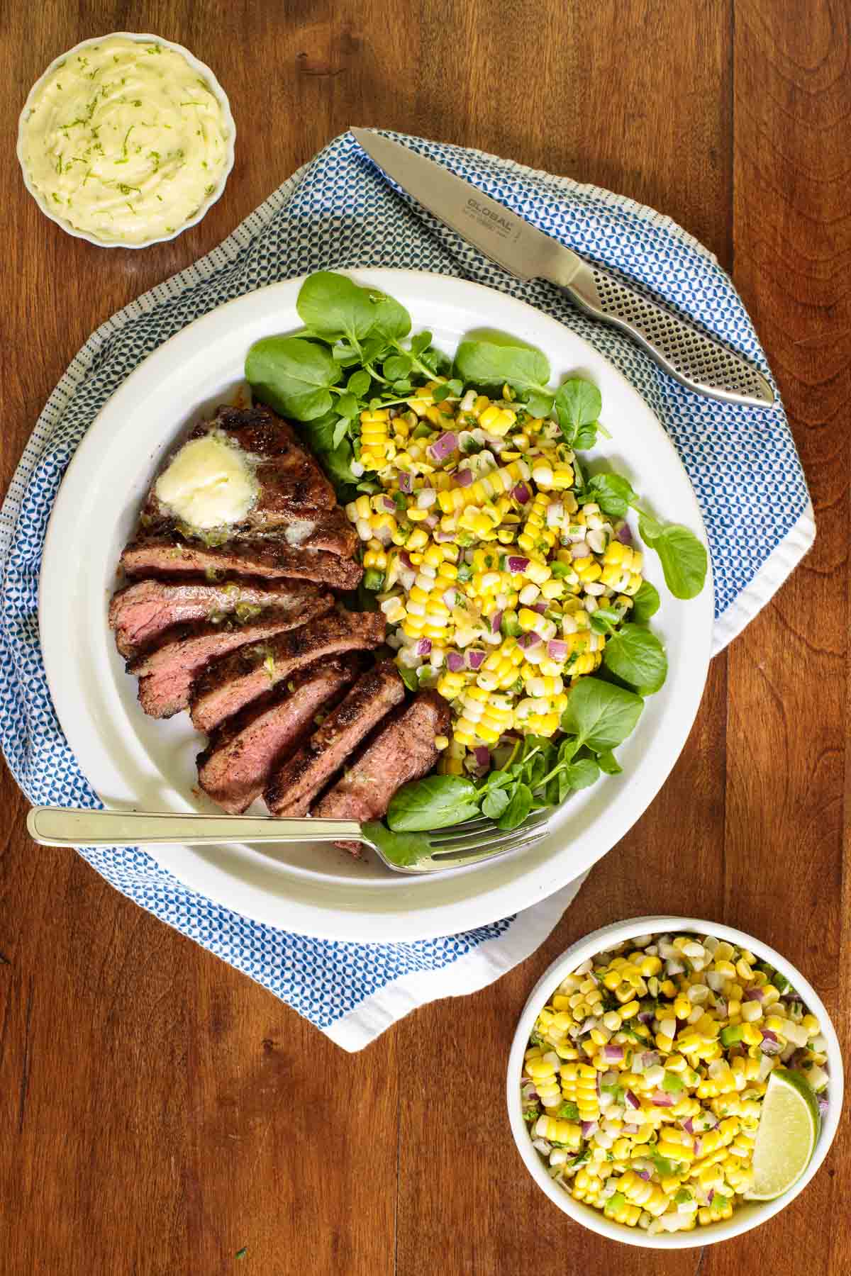 Overhead vertical photo of Perfectly Grilled Steak with corn on a white plate surrounded by a serving of Mexican Street Corn on a wood table.