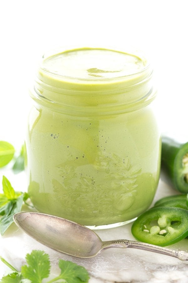 Vertical closeup photo of Peruvian Green Sauce in a glass mason jar surrounded by slices jalapeños and cilantro leaves.