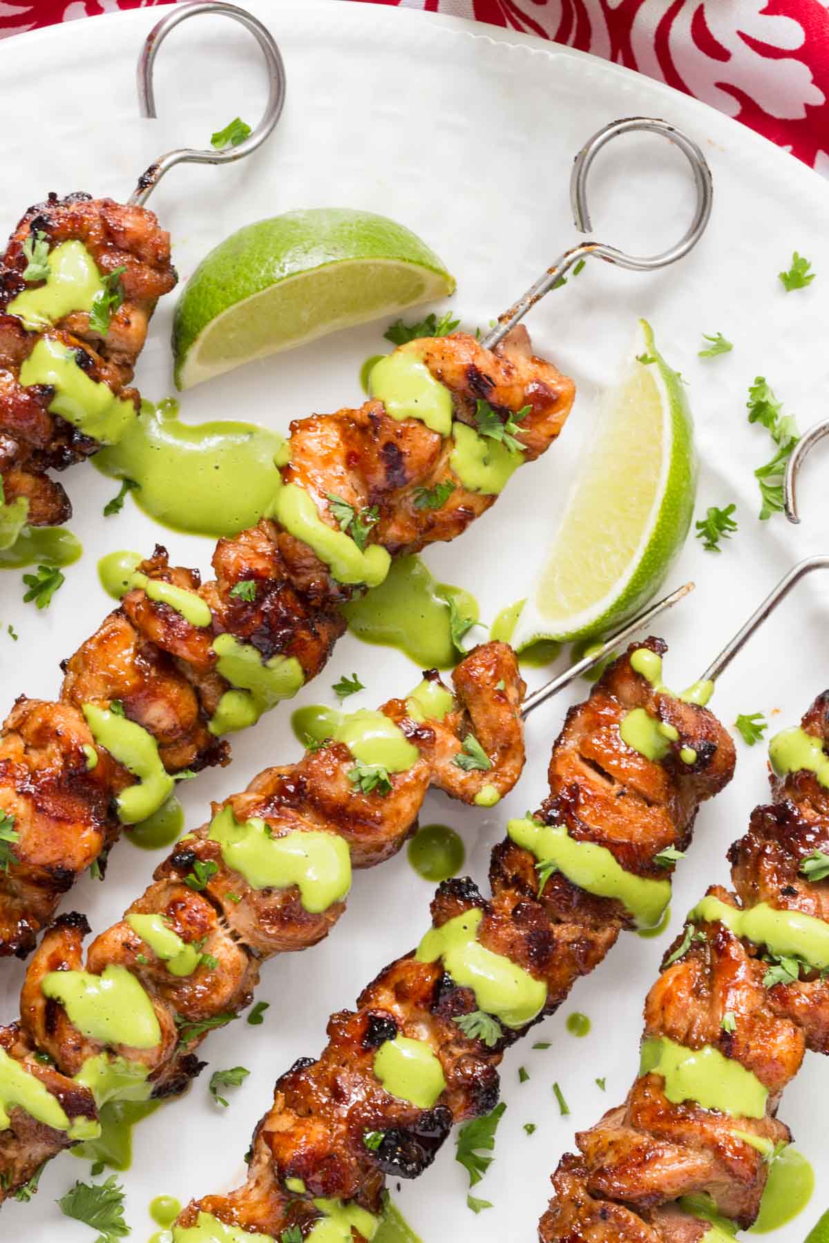 Overhead closeup photo of a white serving plate filled with Peruvian Grilled Chicken Skewers garnished with cilantro, lime wedges and Peruvian Green Sauce.