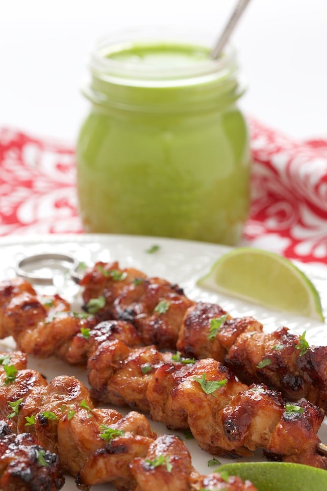 Photo of a white platter filled with Peruvian Grilled Chicken Skewers and a jar of Peruvian Green Sauce in the background.