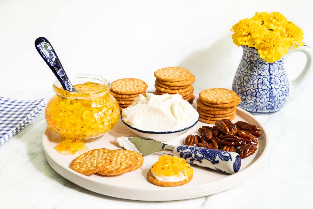 Horizontal photo of a jar of Pineapple Habanero Pepper Jelly on a tray with crackers, cream cheese and candied pecans.
