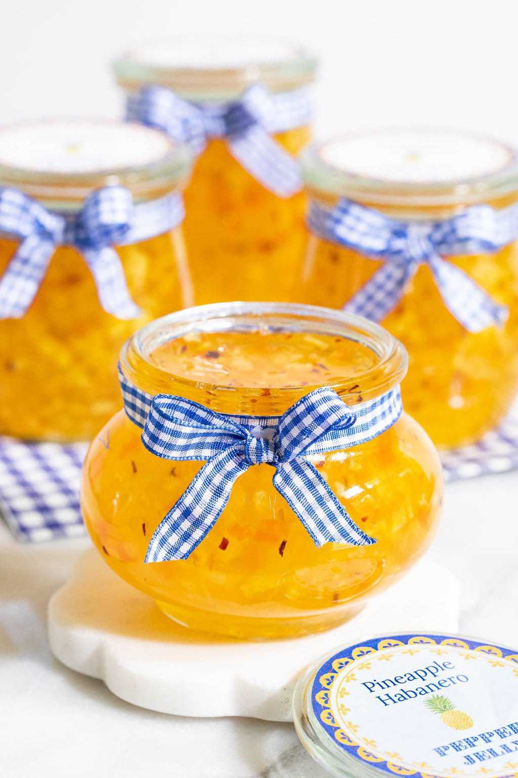 Vertical photo of Pineapple Habanero Pepper Jelly in small glass jars with blue and white ribbon bows and custom labels.