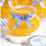Vertical closeup photo of a glass jar of Pineapple Habanero Pepper Jelly decorated with custom labels and blue and white checkered ribbon.