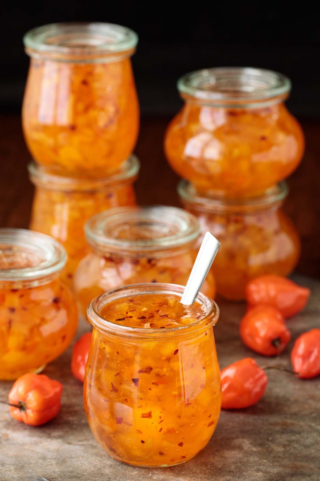 A photo of glass jars filled with Pineapple Habanero Pepper Jelly on a slate table. Jars are stacked up in the background.