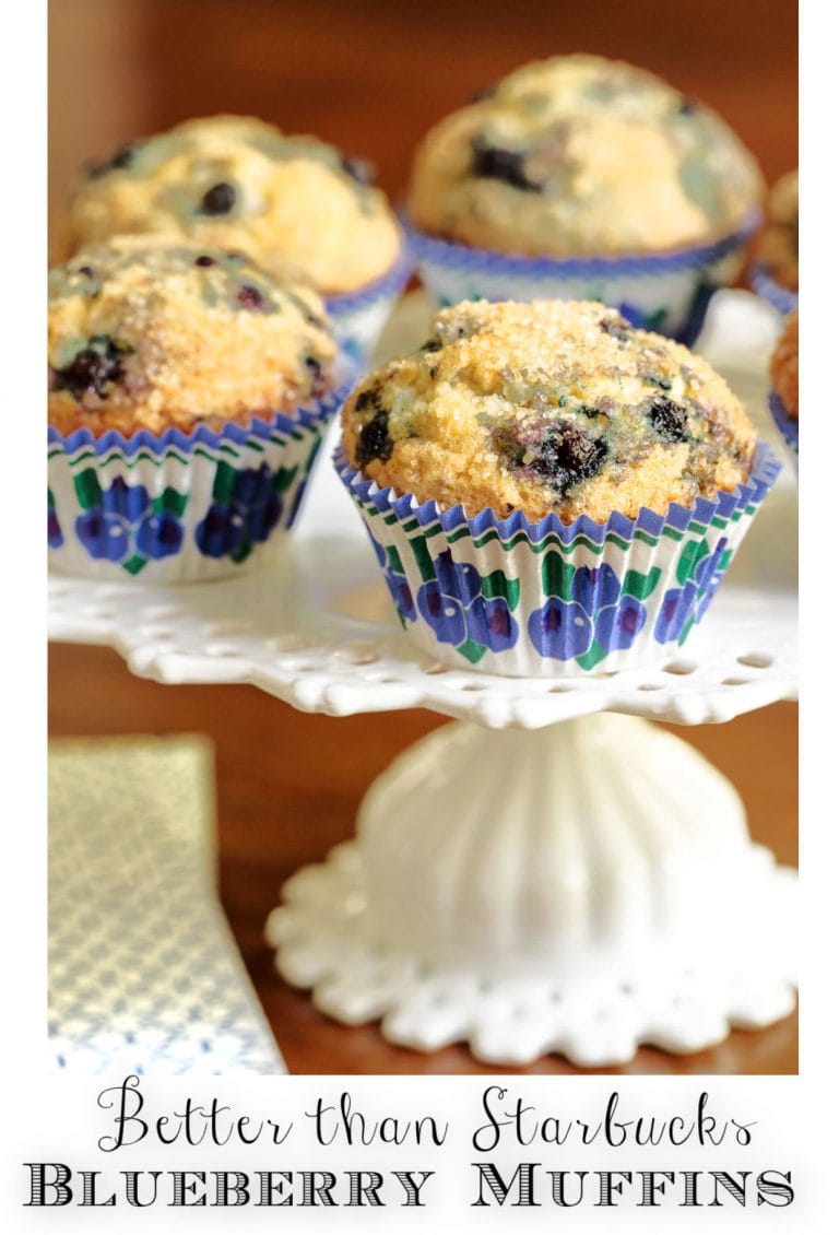 Vertical closeup photo of a batch of Better than Starbucks Blueberry Muffins on a white pedestal serving plate.