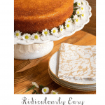 Vertical photo of a Ridiculously Easy Orange Olive Oil Cake on a white scalloped pedestal cake stand and decorated with mini daisies.