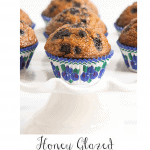 Vertical closeup photo of a batch of Honey-Glazed Blueberry Bran Muffins on a white pedestal plate and resting in white, purple, blue and green cupcake liners.