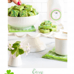 Vertical photo of a bowl of Leprechaun Shortbread Bites on a pedestal display stand with a shamrock plant in the foreground and a quart of Irish milk in the background.