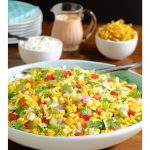 Vertical photo of a Chopped Mexican Street Corn Salad in a white and turquoise serving bowl with dressing and tortilla strips in the background.