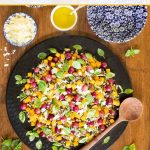 Overhead vertical photo of a platter of Roasted Butternut Squash Farro Salad on a wood table surrounded by fresh mint leaves and bowls of cheese and dressing.