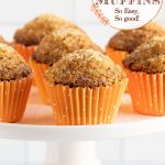 Vertical closeup photo of a batch of Sugar Top Carrot Muffins in orange cupcake liners on a white pedestal display platter.