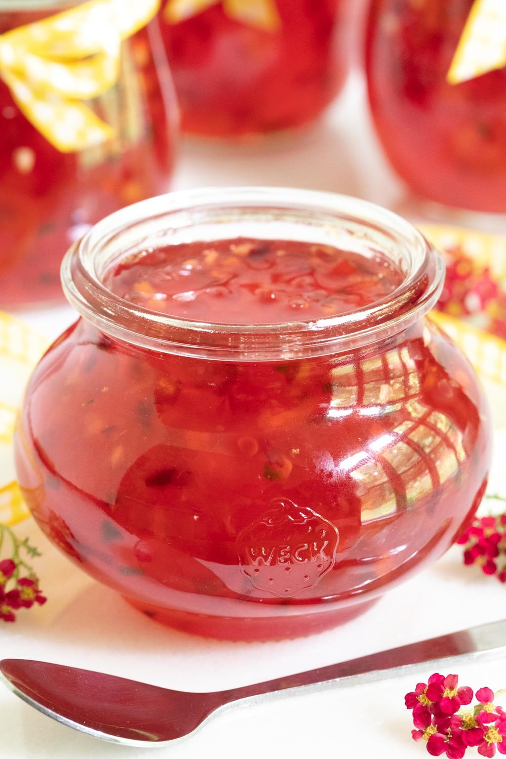 Vertical closeup photo of a glass Weck jar of Plum Ginger Pepper Jelly with additional jars of the jelly in the background.