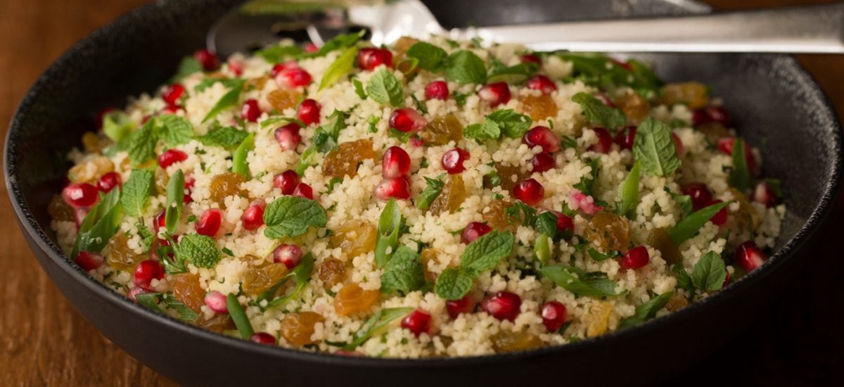 Photo of a black bowl of Pomegranate Parsley Couscous Salad on a wood table.