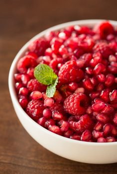 Vertical closeup photo of a bowl of fresh pomegranate seeds and raspberries in a white bowl.