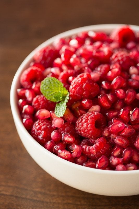 Vertical closeup photo of a bowl of fresh pomegranate seeds and raspberries in a white serving bowl.