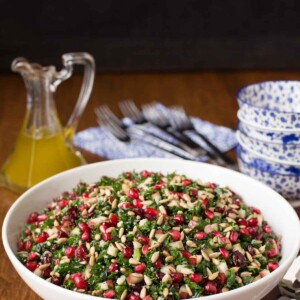 Vertical picture of Pomegranate Pear Salad in a white bowl with blue and white dishes stacked and dressing in a glass cruet in the background