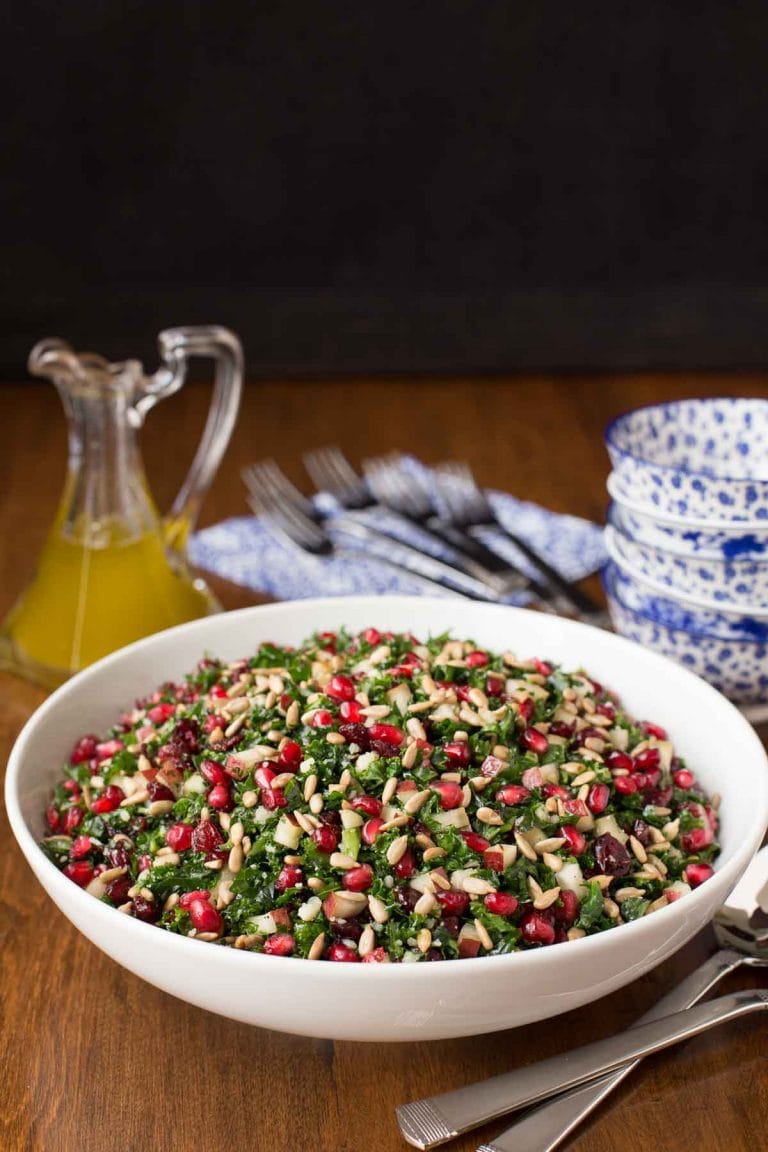 Vertical picture of Pomegranate Pear Salad in a white bowl with blue and white dishes stacked and dressing in a glass cruet in the background