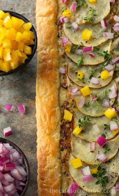 A photo of a Potato Pesto Pepper Galette on a tan stone surface with bowls of diced yellow bell pepper and red onions next to the edge of the crust.