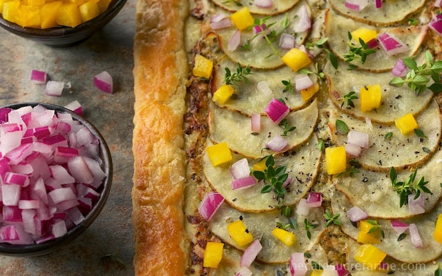 Closeup photo of a Potato Pesto Pepper Galette with bowls of red onions and yellow bell peppers next to the crust.