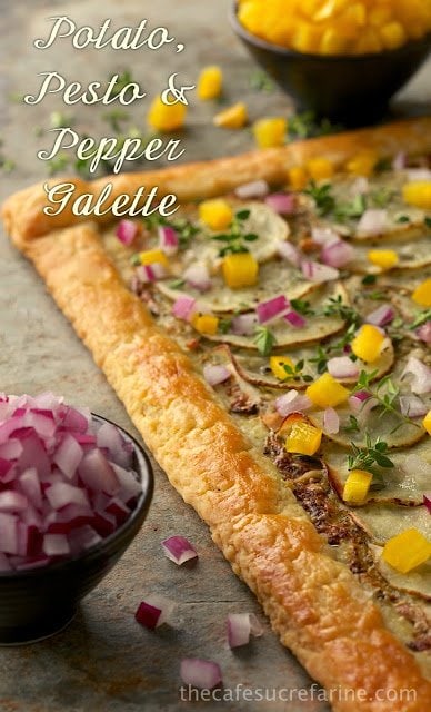 Photo of a Potato Pesto Pepper Galette on a tan stone surface with bowls of diced red onion and yellow bell pepper surrounding the galette.