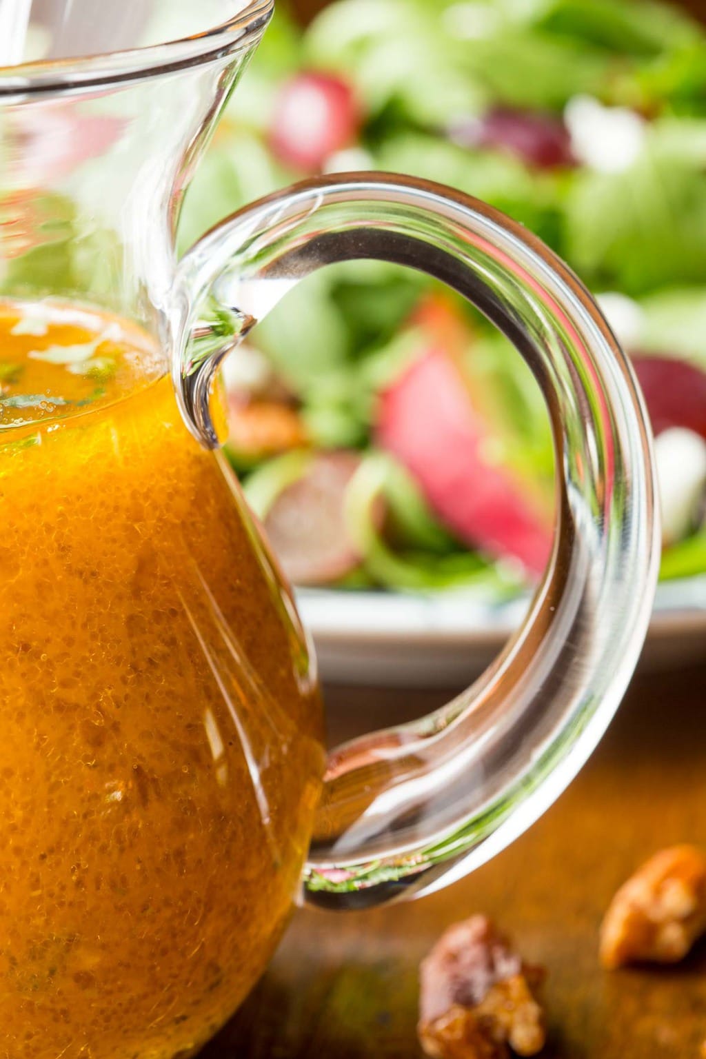Ultra closeup photo of a glass pitcher of Pumpkin Maple Vinaigrette with a fall apple salad in the background.