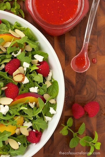 Overhead photo of half a dish of Raspberry Arugula Salad on a wood cutting board with a jar of dressing and a serving spoon next to it.