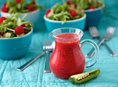 Photo of a pouring jar of Raspberry Honey-Jalapeño Vinaigrette with bowls of arugula and raspberry salad in the background and a slice of a jalapeño pepper next to the jar.