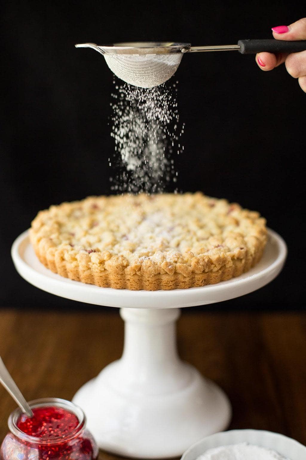 Photo of powdered sugar being sifted on a Raspberry Jam Shortbread Tart. The tart is on a white display pedestal with a jar of raspberry jam and a dish of powdered sugar in the foreground.