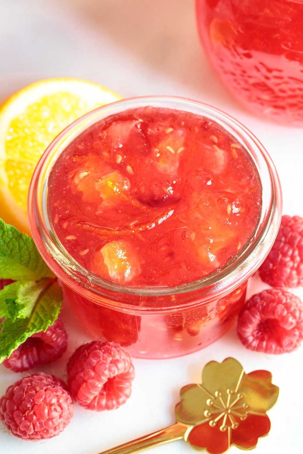 Vertical extreme closeup photo of a glass jar of Raspberry Meyer Lemon Marmalade surrounded by fresh raspberries and lemons.