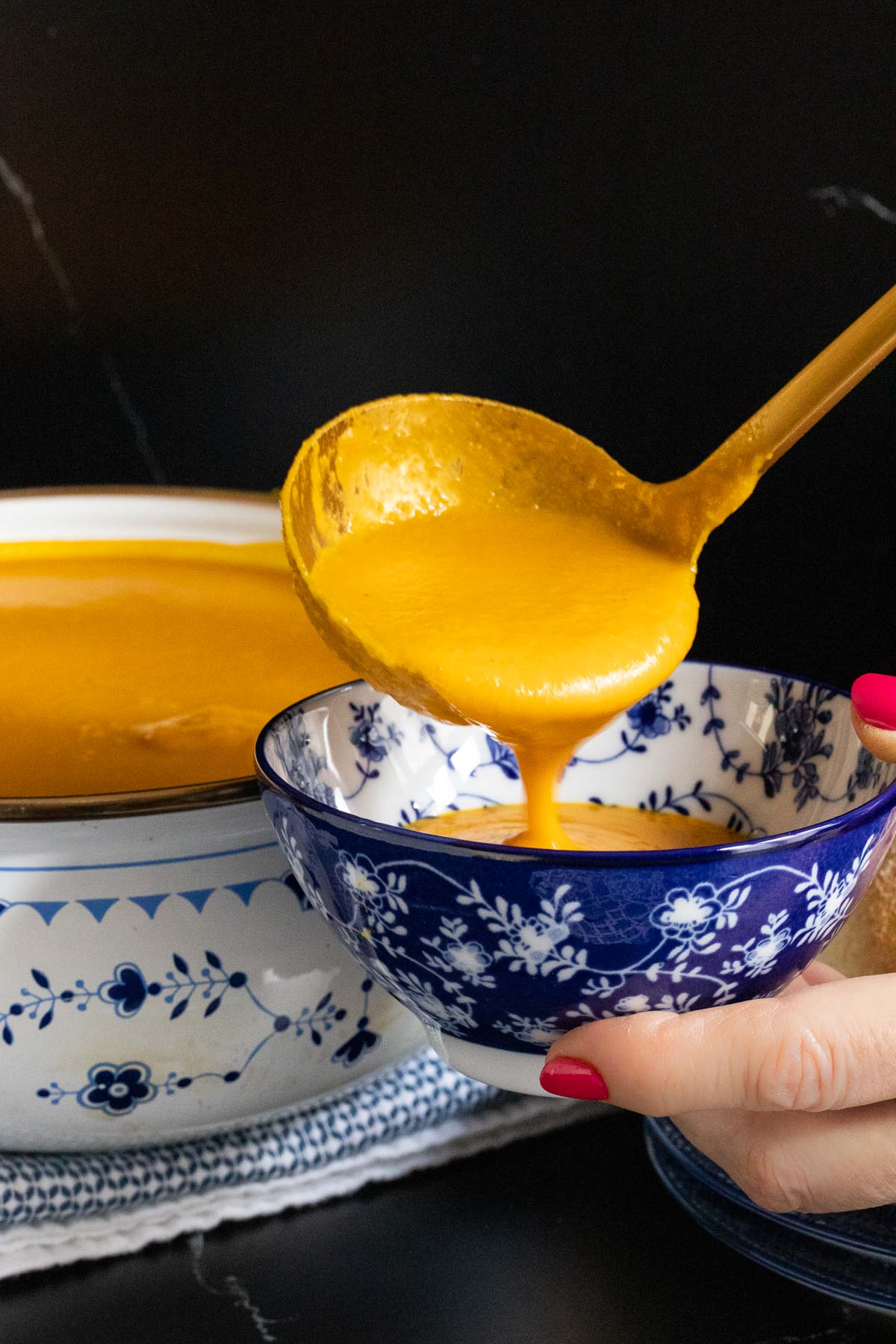 Vertical closeup photo of a person pouring Red Lentil Carrot Ginger Soup into a blue and white patterned individual serving bowl.