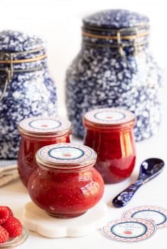 Vertical picture of Red Raspberry Freezer Jam in glass jars with gift labels with white a blue pottery in the background