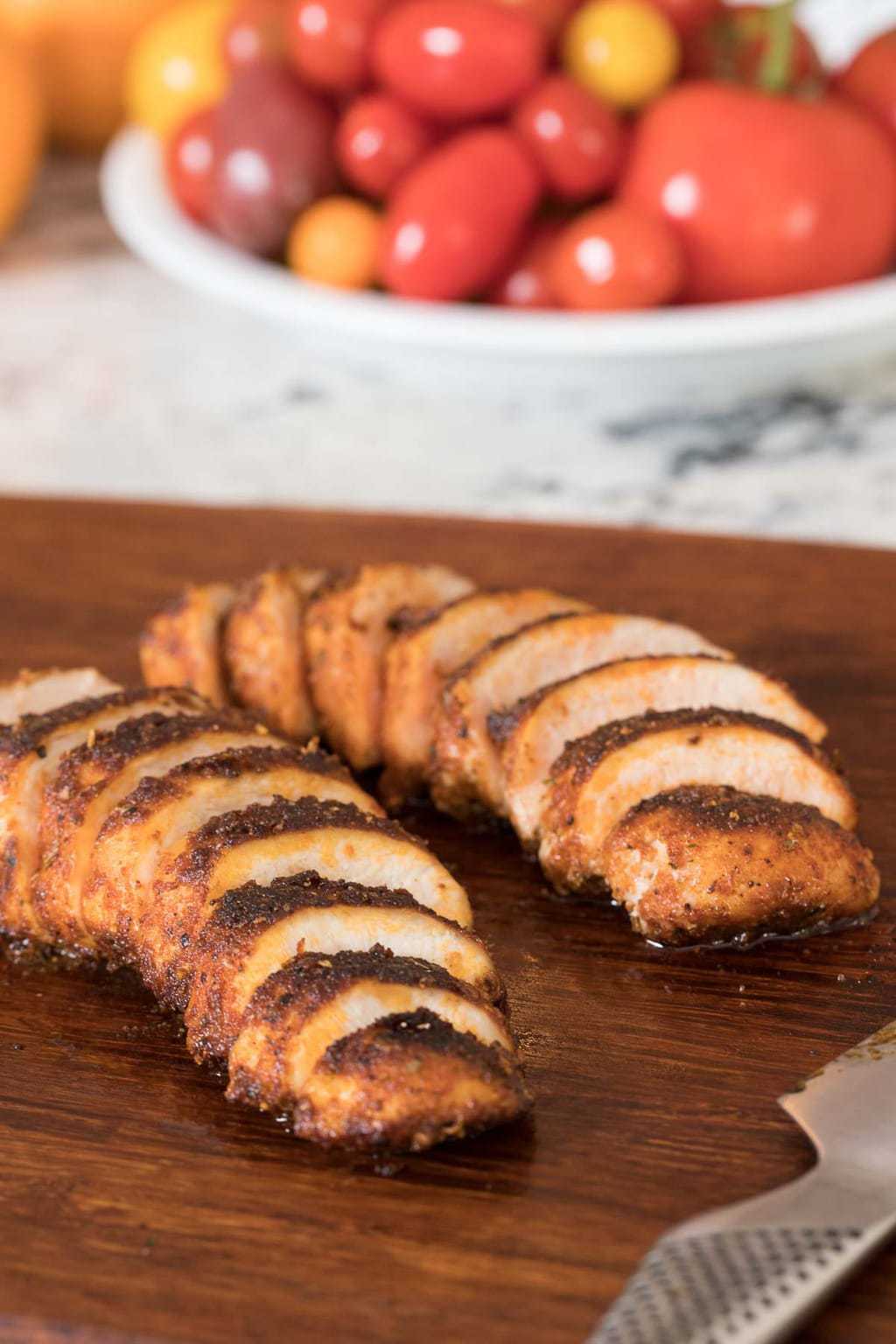 Photo of Restaurant Style Sautéed Chicken Breasts sliced on a cutting board. A plate of fresh tomatoes is in the background.