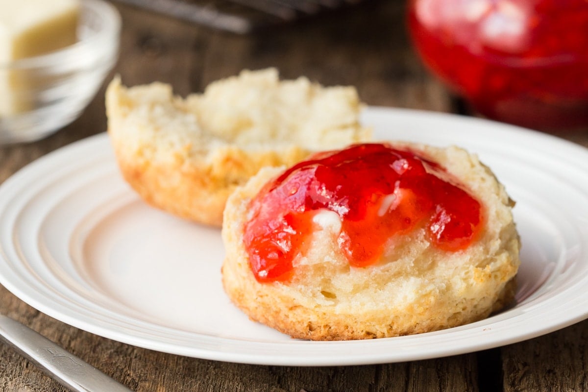 Horizontal closeup photo of a Ridiculously Easy Buttermilk Biscuit on a white serving plate with melted butter and strawberry jam on top.