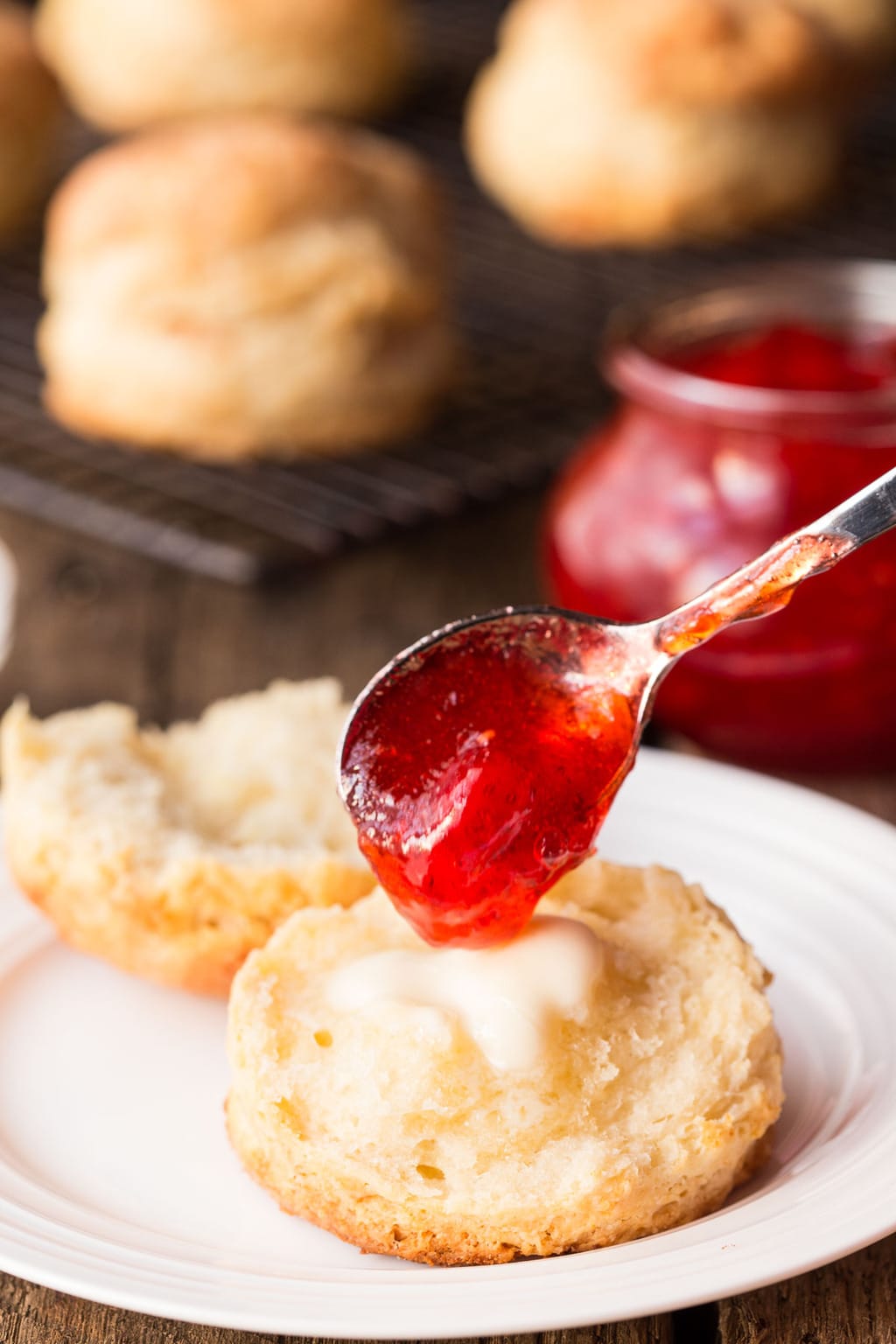 Vertical photo of Ridiculously Easy Buttermilk Biscuits topped with butter and a spoonful of strawberry jam. More biscuits are on a metal cooling rack in the background.