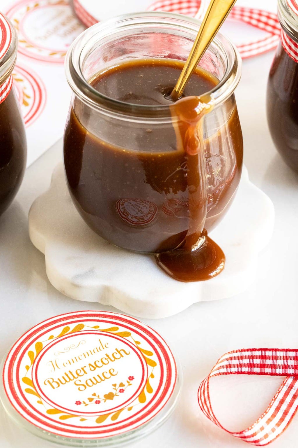 Vertical photo of a glass jar of Ridiculously Easy Butterscotch Sauce with custom gift labels in the foreground and background.