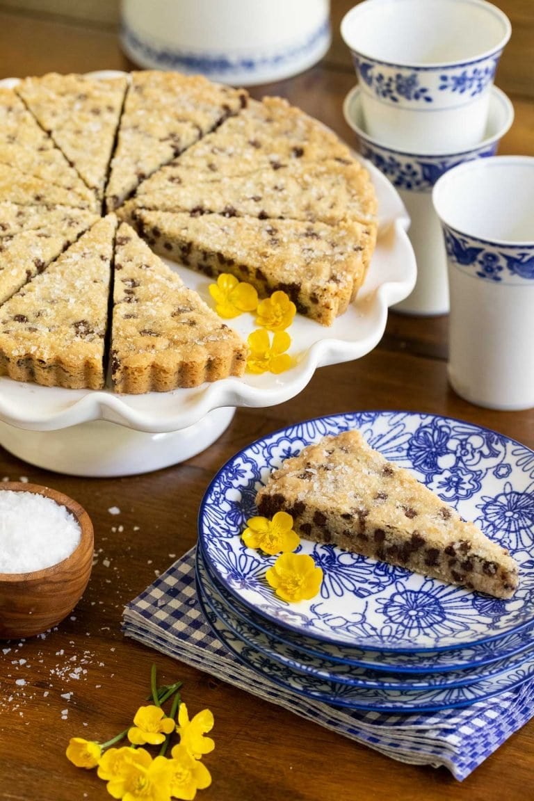 Vertical picture of chocolate chip shortbread cut into wedges on a cake stand with blue and white plates