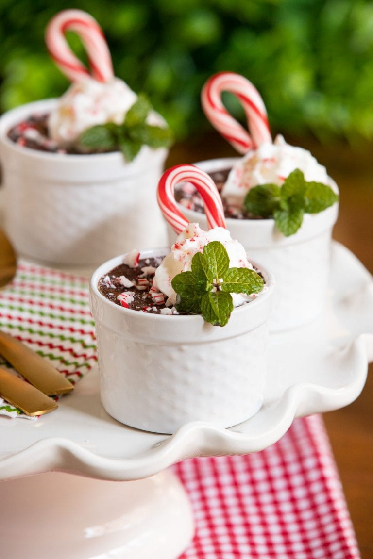 Vertical photo of cups of Ridiculously Easy Chocolate Peppermint Pots de Crème on a scalloped serving platter. The cups are garnished with whipped cream, fresh mint leaves and candy canes.