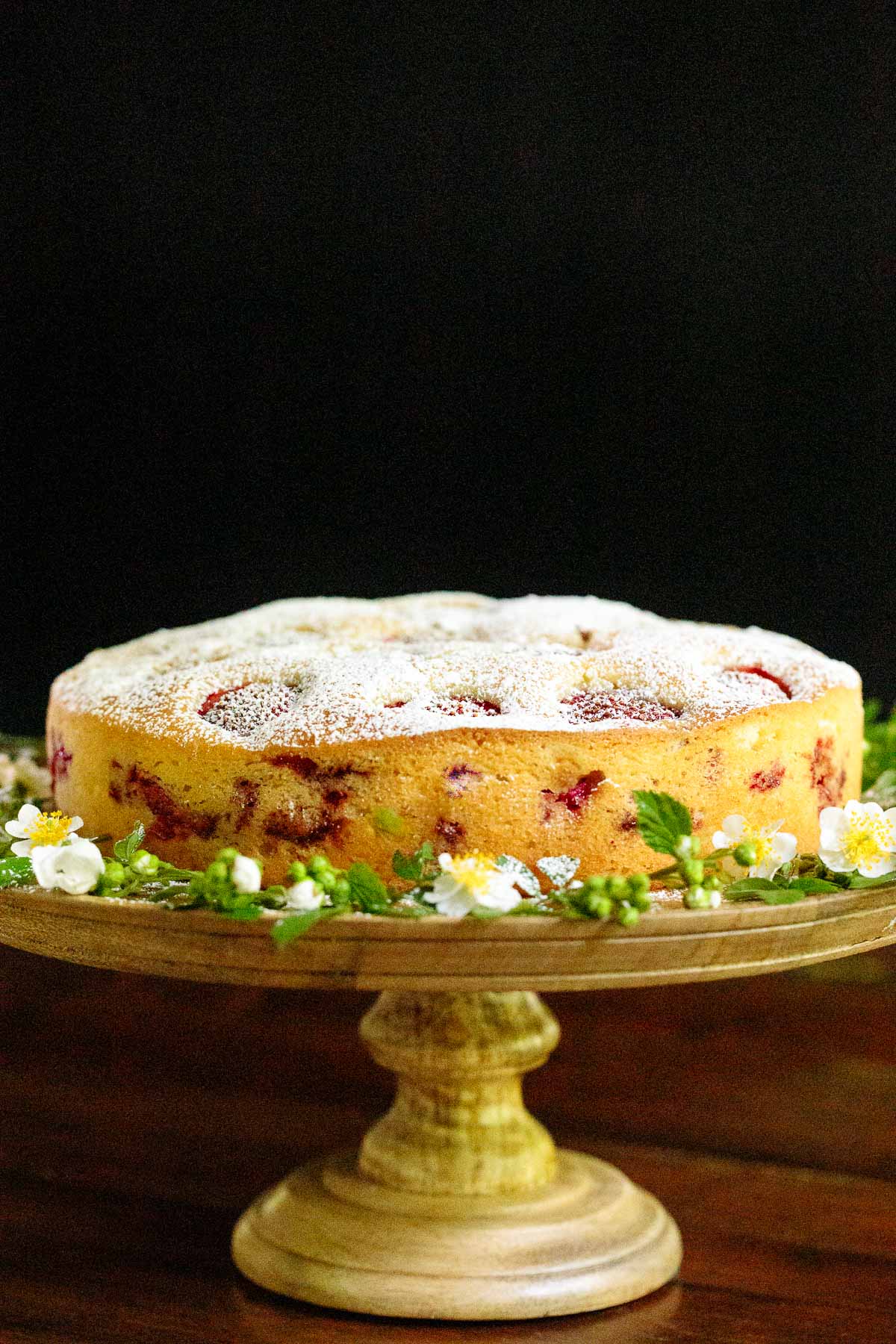 Vertical photo of a Ridiculously Easy Fresh Strawberry Cake on a rustic wood pedestal cake stand against a black background.
