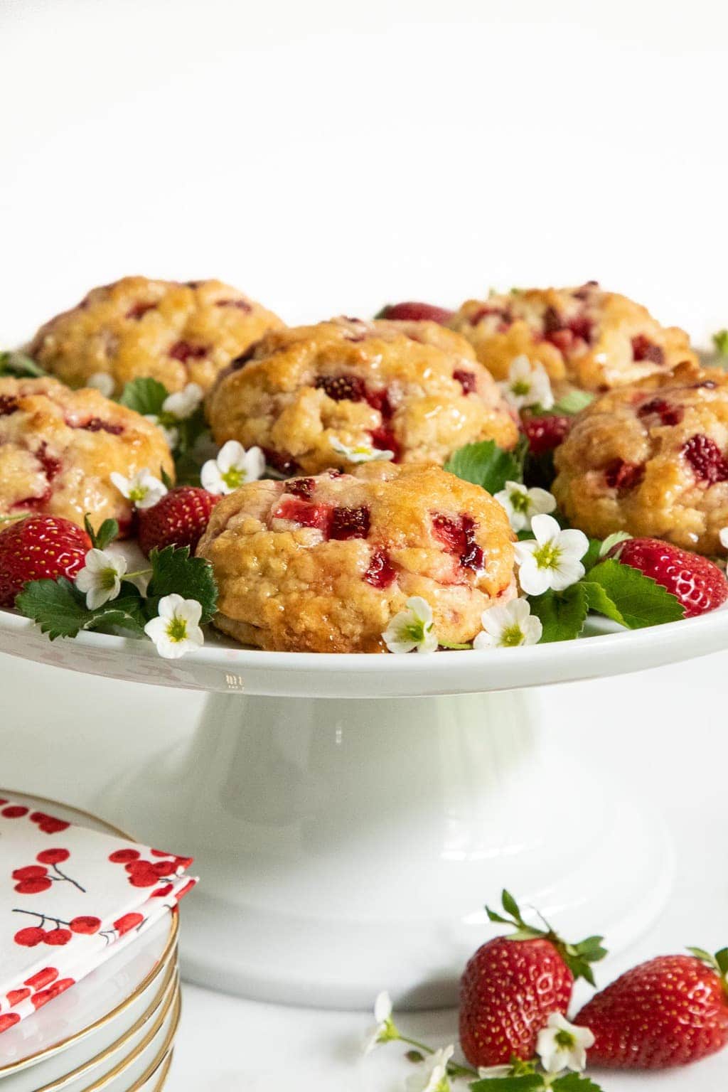 Vertical photo of a white pedestal platter of Ridiculously Easy Fresh Strawberry Scones decorated with strawberry leaves and flowers.