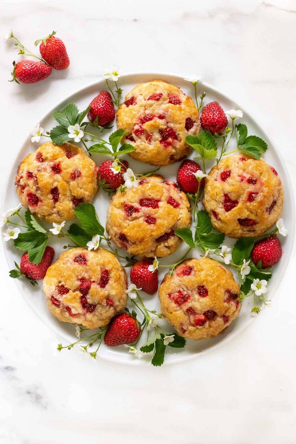 Vertical overhead photo of a white round platter of Ridiculously Easy Fresh Strawberry Scones garnished with strawberry leaves and flowers.