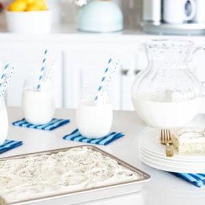 Horizontal photo of a Ridiculously Easy Lemon Poppy Seed Sheet Cake on a kitchen countertop with glasses and a pitcher of milk in the background.
