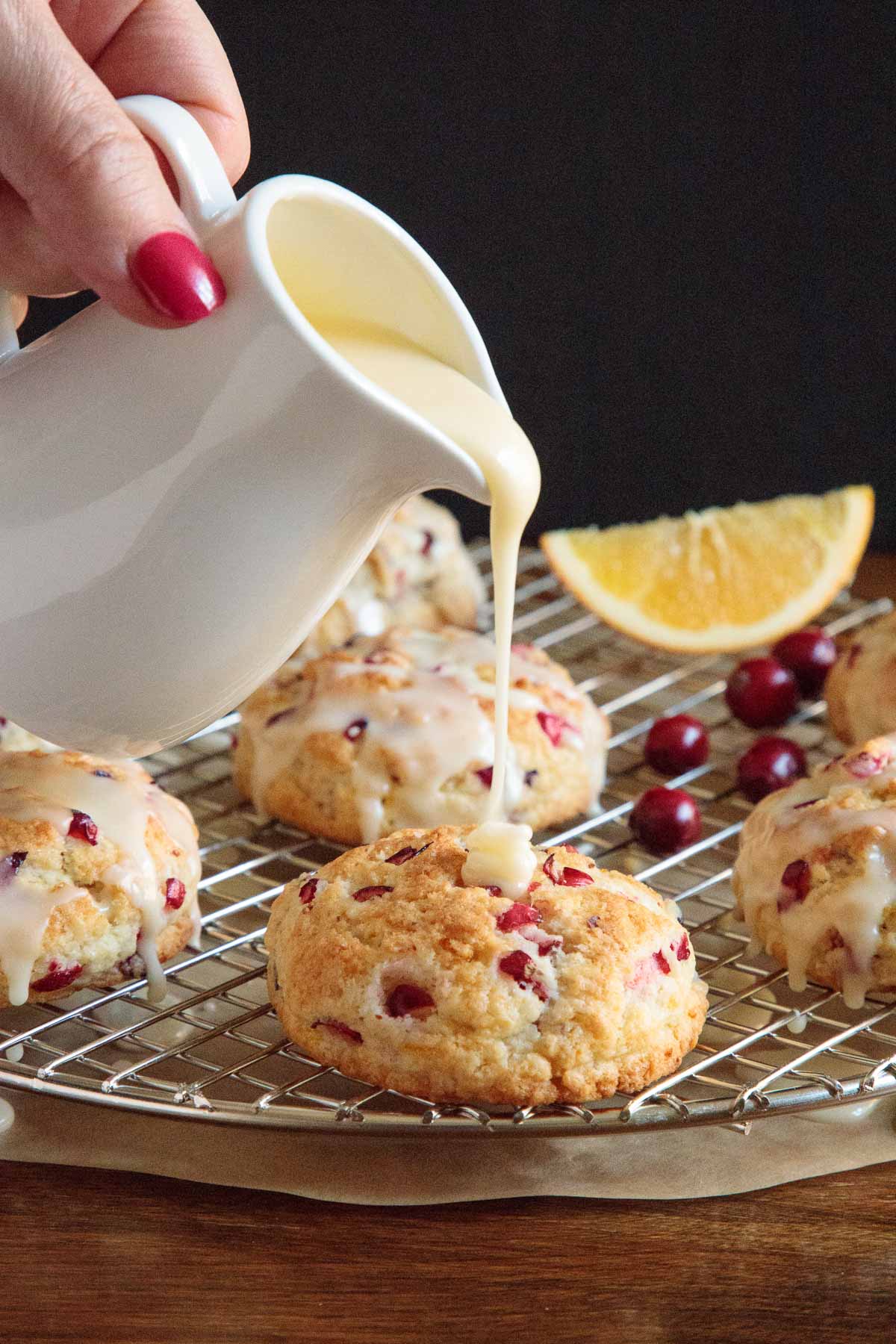 Vertical closeup photo of a hand pouring orange icing over a batch of Ridiculously Easy Orange Cranberry Scones on a cooling rack.