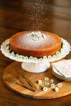 Vertical photo of a Ridiculously Easy Orange Olive Oil Cake on a scalloped pedestal cake stand and decorated with mini daisies.