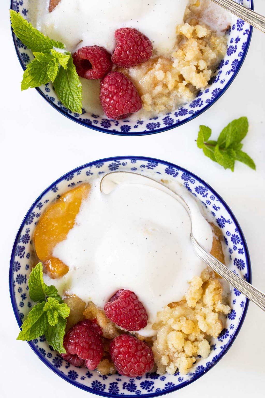 Overhead vertical photo of two blue and white bowls filled with Ridiculously Easy Peach Crumble with fresh raspberries and whipped cream.
