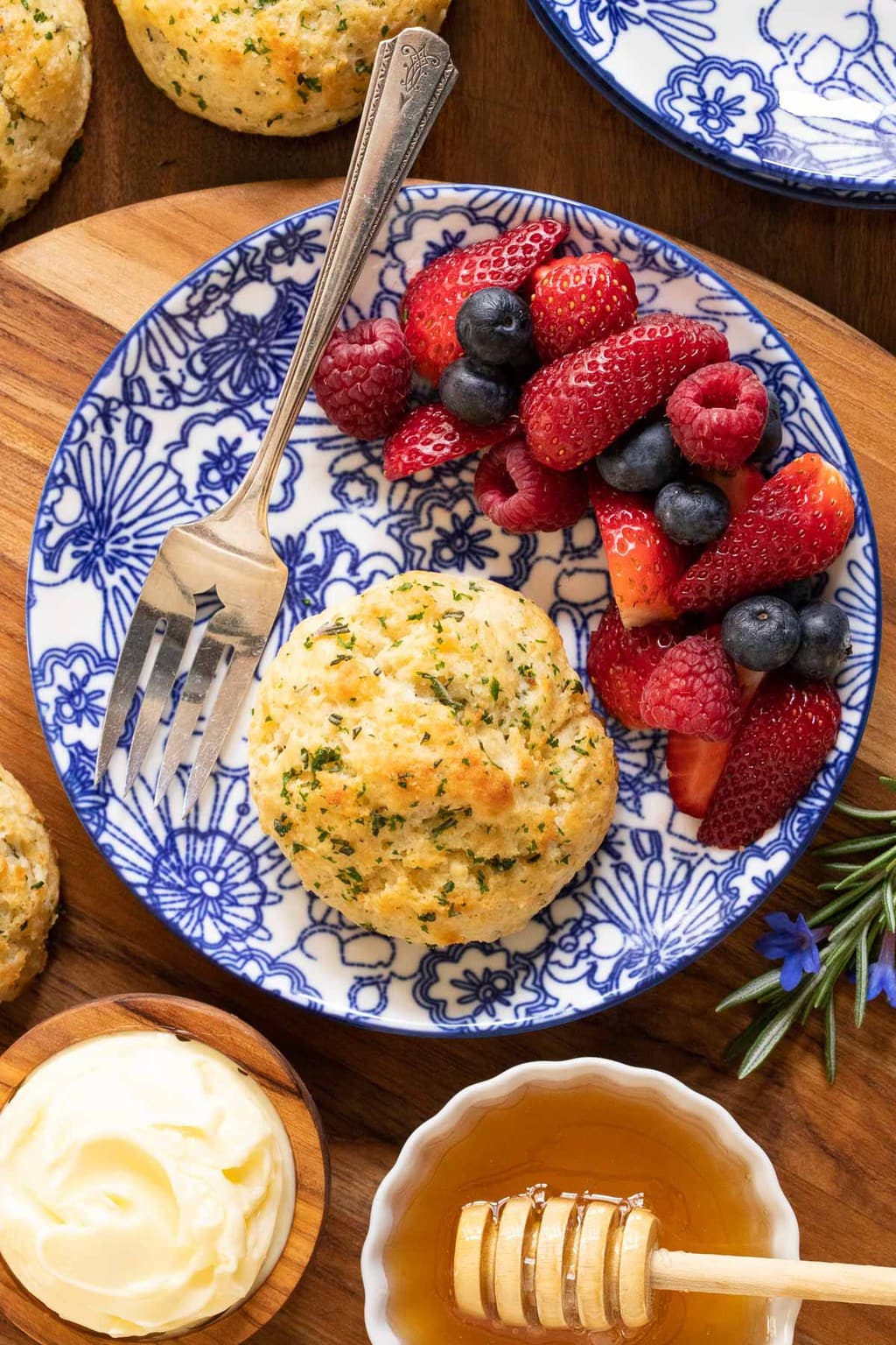 Vertical overhead photo of Ridiculously Easy Rosemary Parmesan Biscuits on a blue and white patterned plate with fresh berries, honey and butter.
