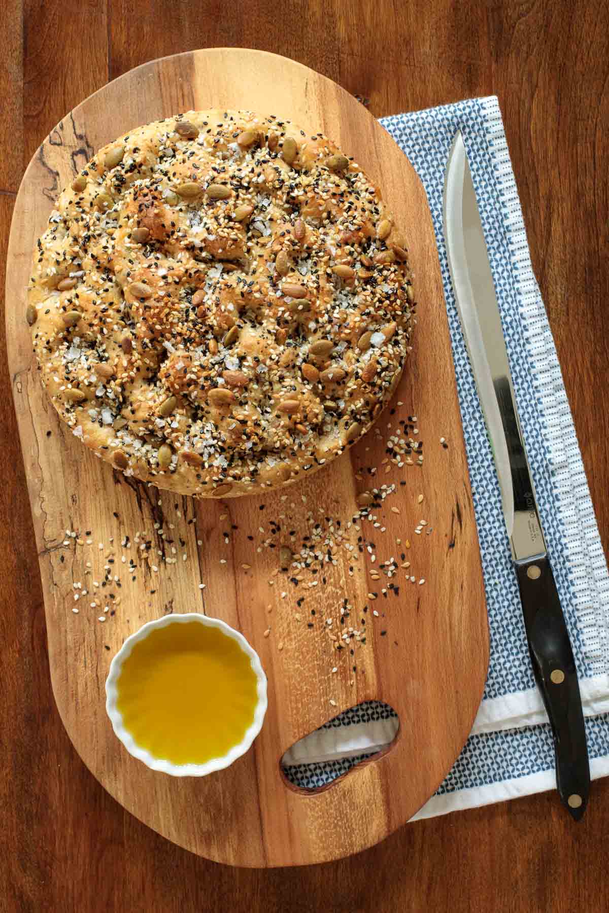 Overhead photo of a loaf of Ridiculously Easy Seeded Focaccia Bread on a wood cutting board with a bread knife and a blue and white patterned towel next to the board.